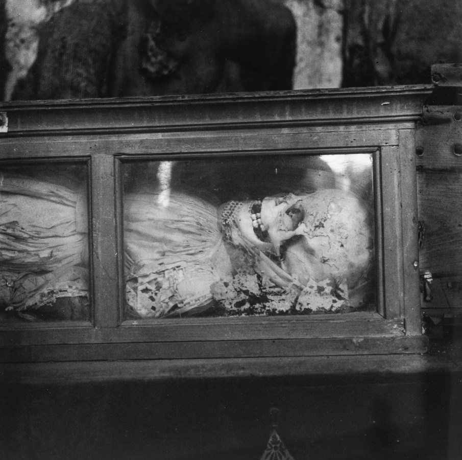Black-and-white photograph of a skeleton in a lace dress lying in a glass-fronted box