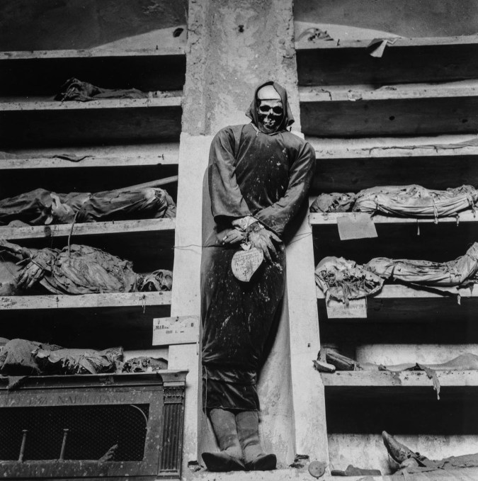 Black-and-white photograph of a hooded preserved body propped up amid other skeletons in compartments