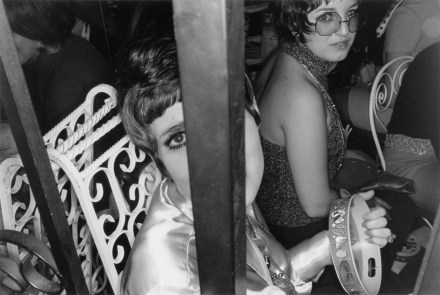 Black and white photograph of a seated young woman holding a tambourine looking at camera through iron gate