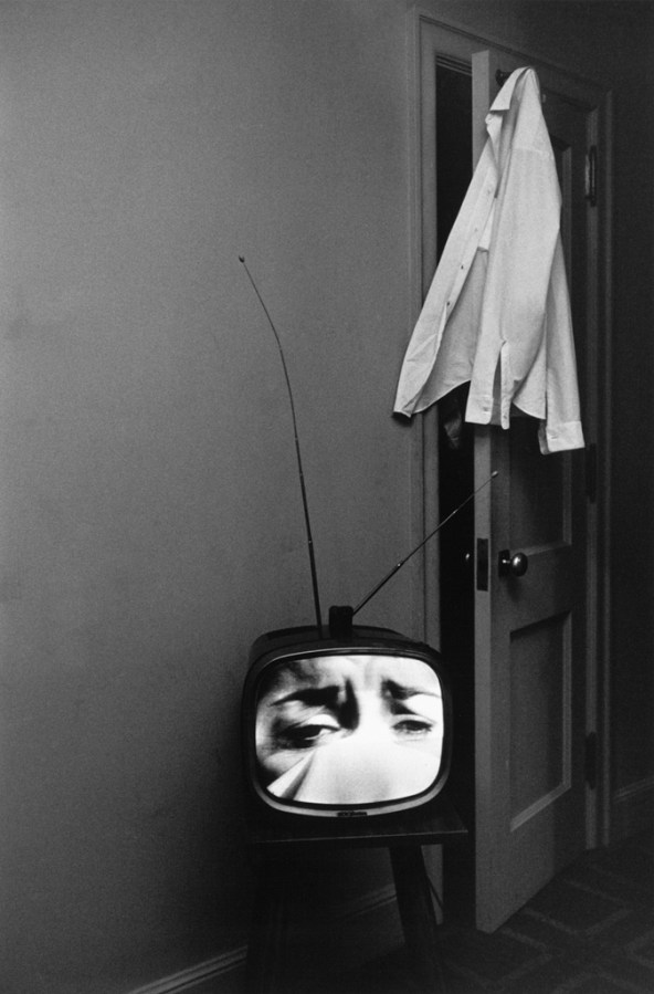 Black-and-white photograph of a television with a crying face on screen and a shirt hanging from an open door