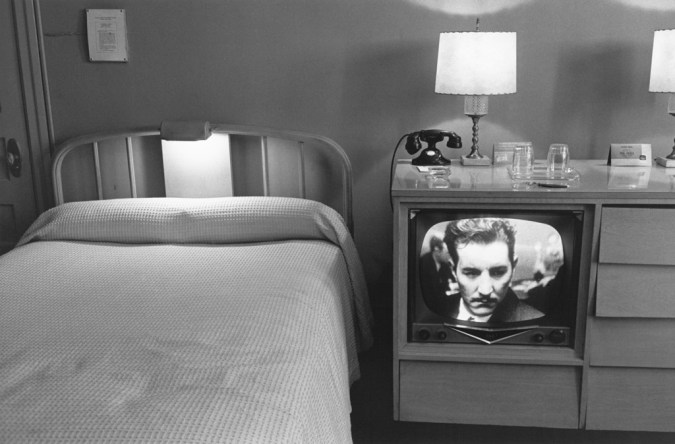 Black-and-white photograph of a neatly made bed and a television with a mans face on screen