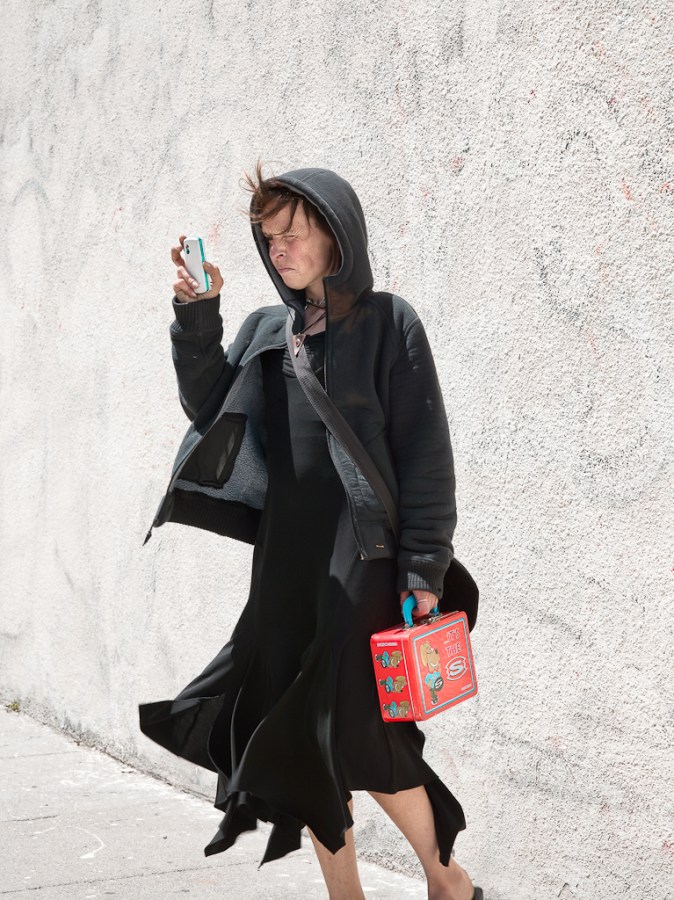Color photograph of a woman dressed all in black peering at a cellphone and holding a red lunchbox