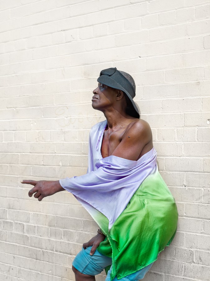 Color photograph of a woman in a lilac and green top leaning against a wall and pointing off-camera