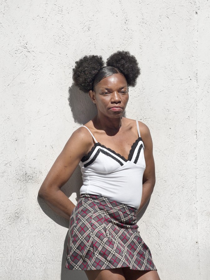 Color photograph of a woman with her hair in two buns leaning against a blank white wall