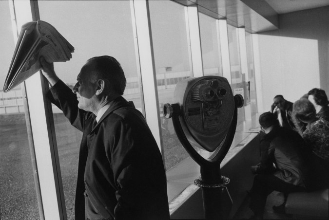 Black-and-white photograph of man looking out a sunny window shielding face with newspaper