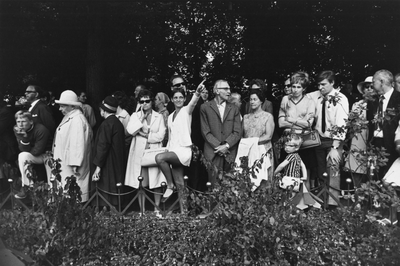 Black and white photograph of people standing in a line along a garden bed. One women in the center points up and to the right.