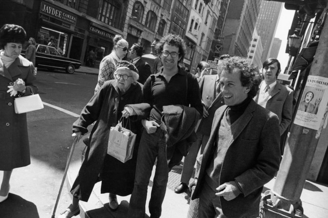 Black-and-white photograph of two smiling men with curly hair accompanying an elderly woman about to cross the street