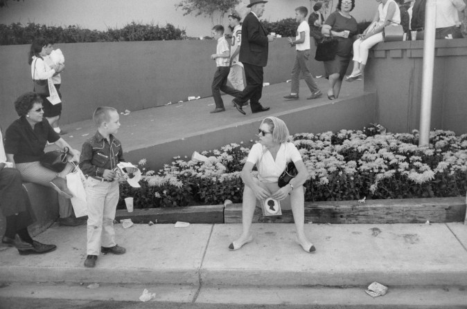 Black-and-white photograph of a woman sitting on a curb holding a cameo portrait and looking at a young boy