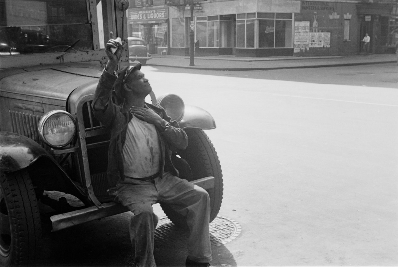 Black-and-white photograph of a man sitting on the front bumper of a vehicle with his arm raised