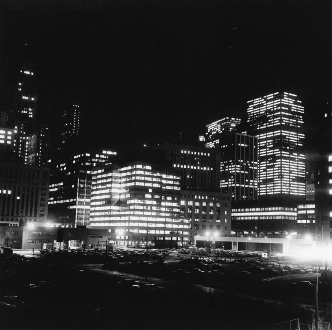 Black-and-white photograph of city buildings lit up from inside standing above a parking lot