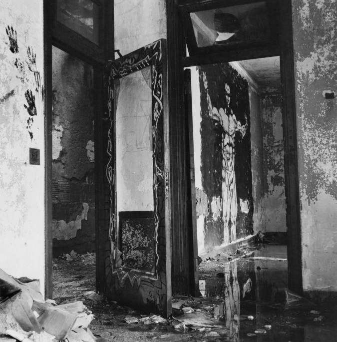 Black-and-white photograph of a derelict interior with a muscular man painted on a wall beyond a doorway