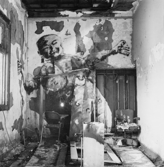 Black-and-white photograph of a derelict interior with a man painted on the wall