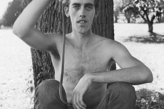 Black and white photograph of a shirtless male figure seating at the base of a tree and holding a snake