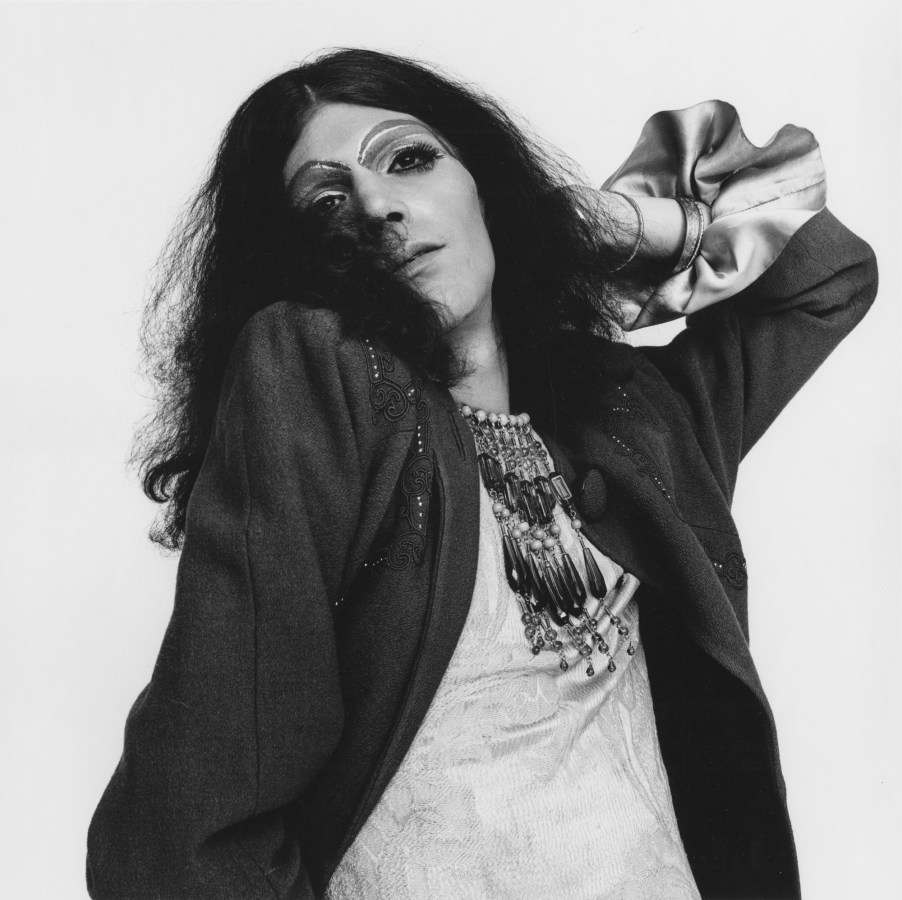 Black-and-white photograph of a person with long wiry black hair and heavy jewelry and make up with one hand behind their head