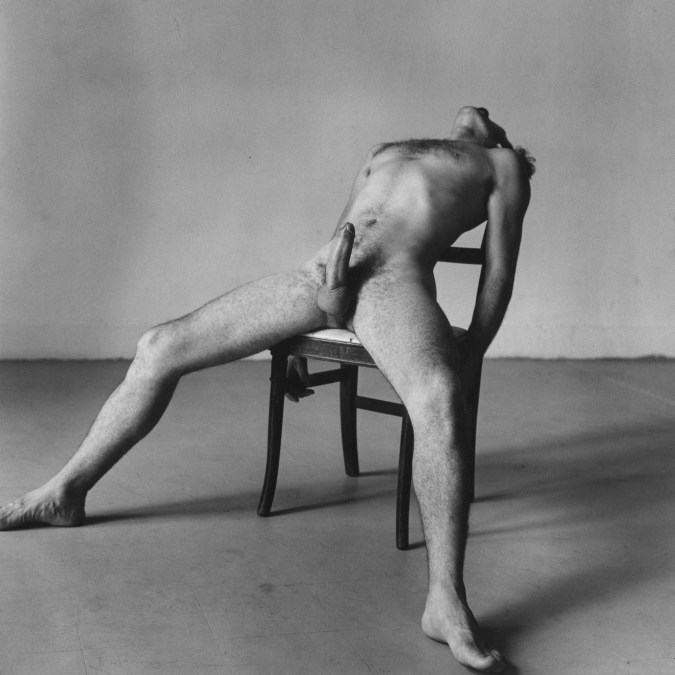 Black-and-white photograph of a seated nude man with an erection bending backward in his chair