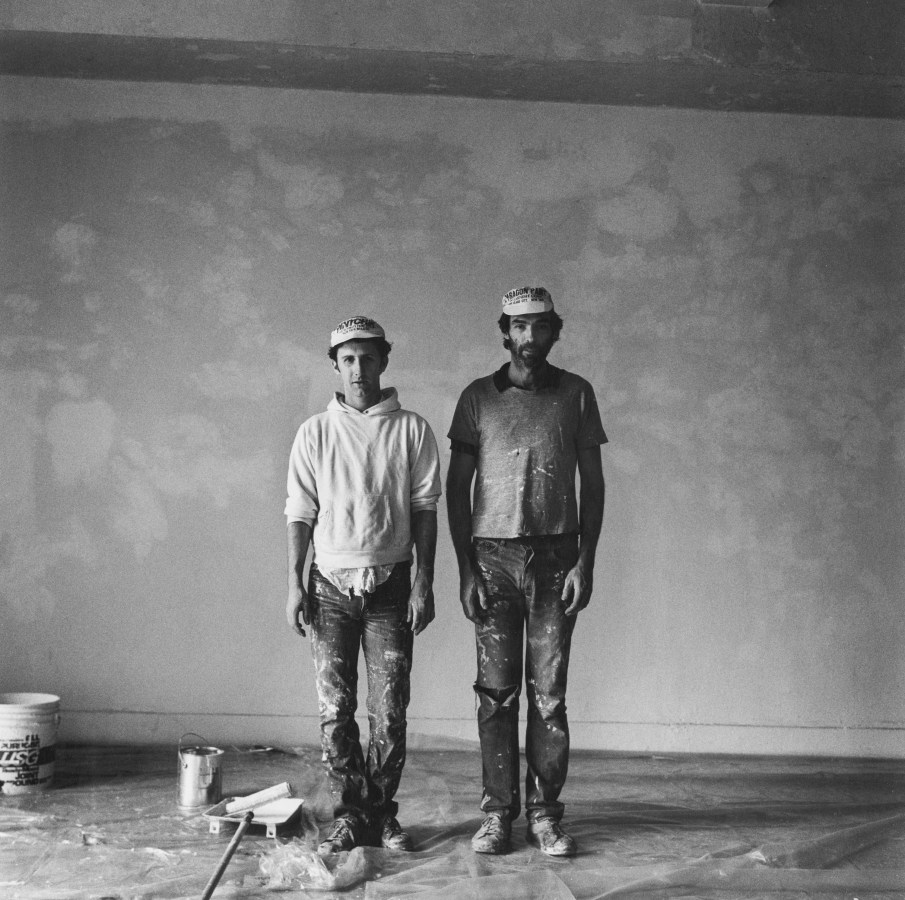 Black and white photograph of two figures wearing hats standing side by side in front of painted wall