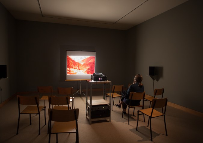 A single person in a darkened room with chairs, a slide projector, and an image of a landscape projected on the screen