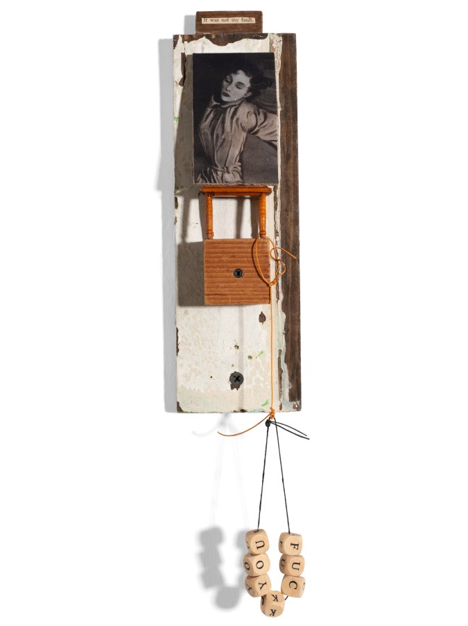 Color image of a mixed media sculpture made up of scrap wood pieces along with a black and white image of a female figure resting with text above reading 'it was not my fault' and wooden block pieces spelling out 'fuck you' on a white wall