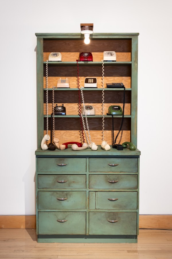 Color image of nine rotary phones on an aged green armoire in a white walled gallery
