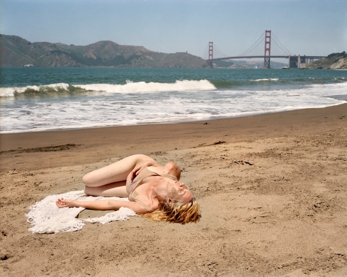 Color photograph of a blonde woman reclined on the sand with the ocean and Golden Gate Bridge in the background.