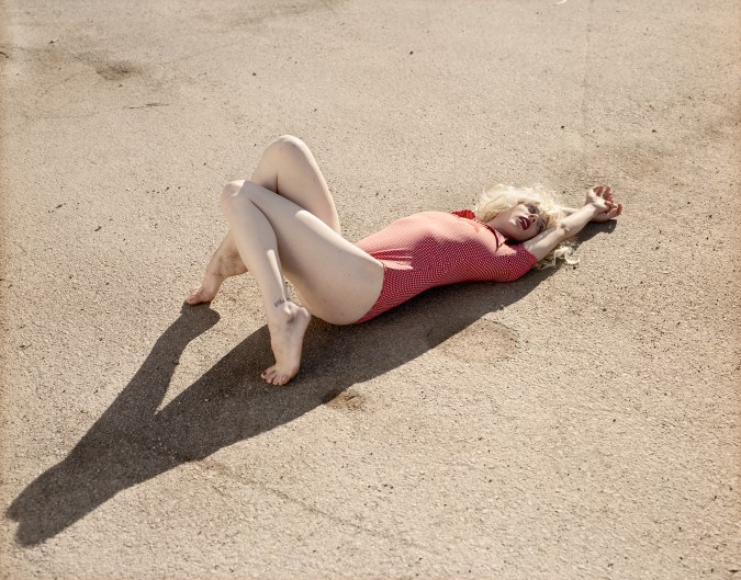 Color photograph of a woman in a red bodysuit lying down on asphalt