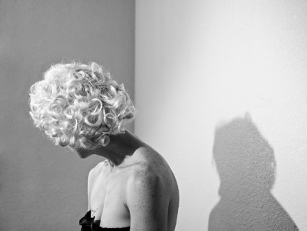 Black and white photograph of a woman in a blonde wig, turned away from the camera