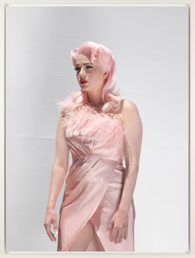 Color photograph of a figure with pink hair wearing a pink dress with a tear coming down their face in front of a white wall framed in white