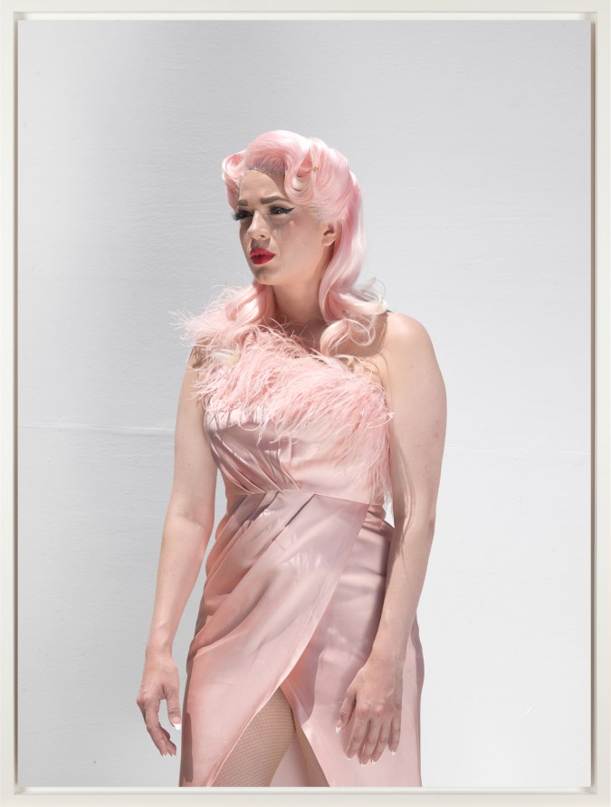 Color photograph of a figure with pink hair wearing a pink dress with a tear coming down their face in front of a white wall framed in white