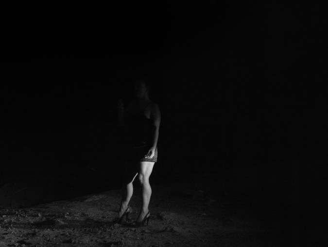 Black-and-white photograph of a woman in high heels standing in the dark with only her legs and arm illuminated