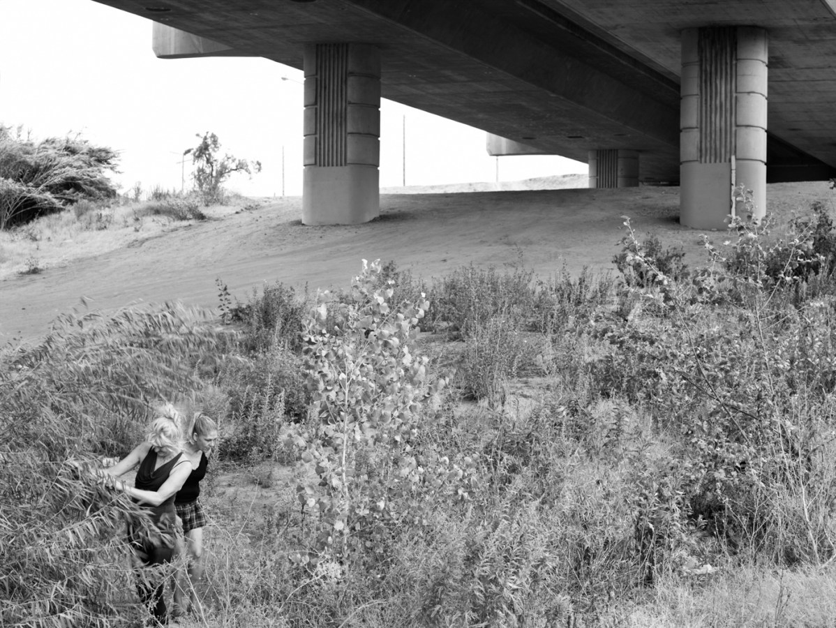 Black-and-white photograph of two women standing in long grass under a concrete bridge