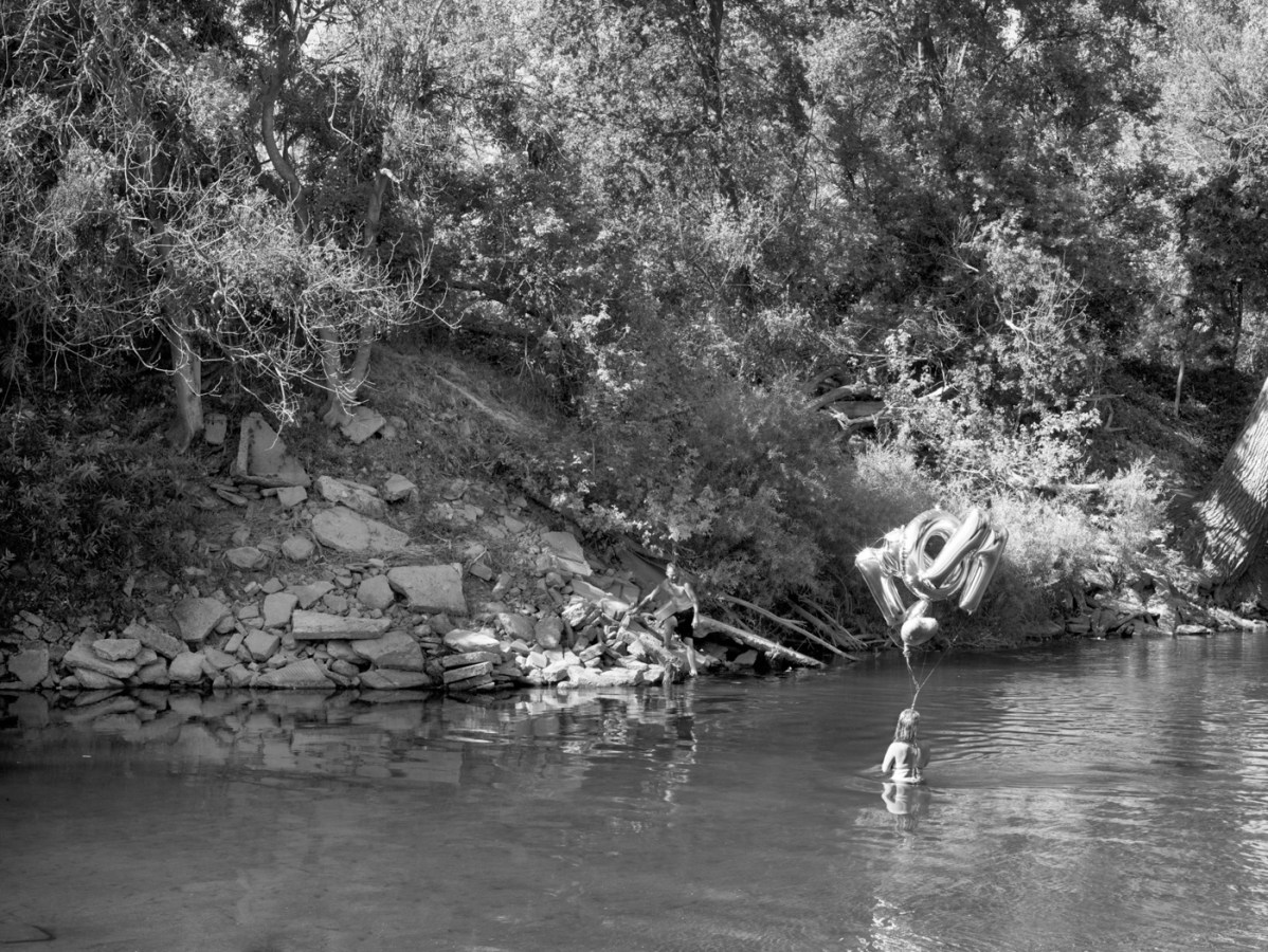 Black-and-white photograph of a man on on a rocky overgrown riverbank watching a woman standing in the water holding foil balloons