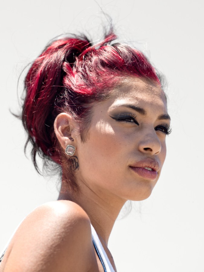 Color photographic portrait of a young woman with dyed red hair in a bun in front of a blank white wall