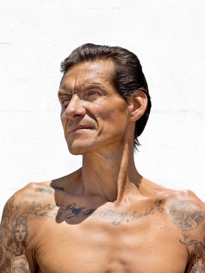 Color photographic portrait of a shirtless man with tattoos across his collarbone against a blank white wall