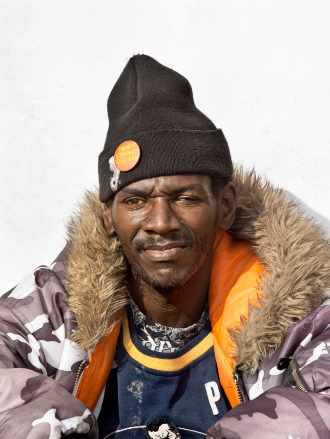 Color photographic portrait of a man in a faux-fur trimmed jacket and beanie hat standing in front of a blank white wall