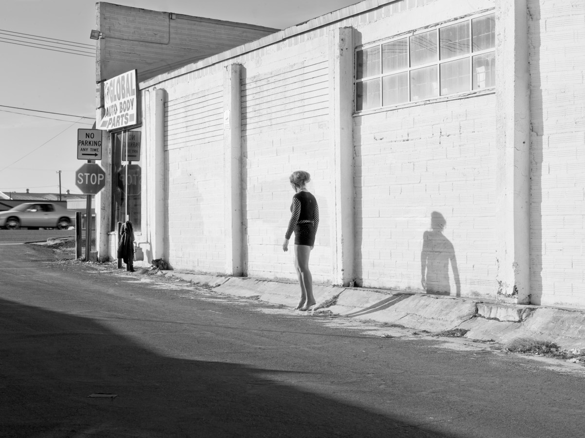 Black-and-white photograph of a woman walking by a low white building at an intersection
