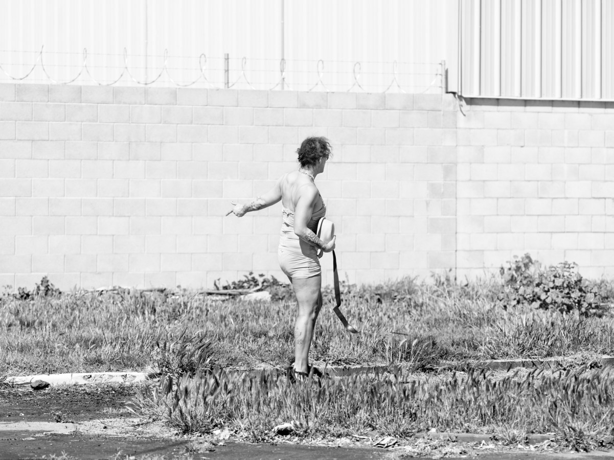 Black-and-white photograph of a woman in an overgrown lot pointing behind her