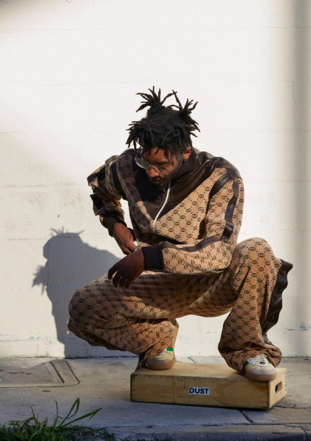 Color photograph of a man in a tracksuit crouching on a wooden box in front of a blank white wall