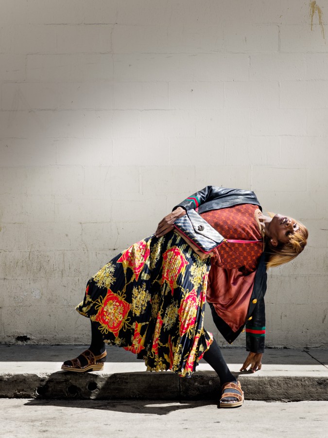 Color photograph of a man in a long pleated skirt and leather jacket bending over sideways
