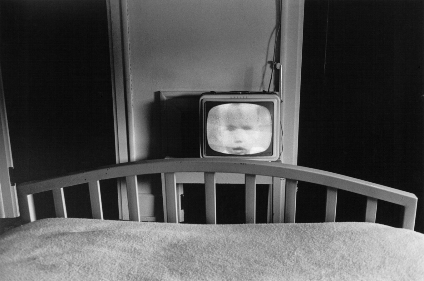 Black-and-white photograph of the bottom edge of a bed and a television with a childs face on screen