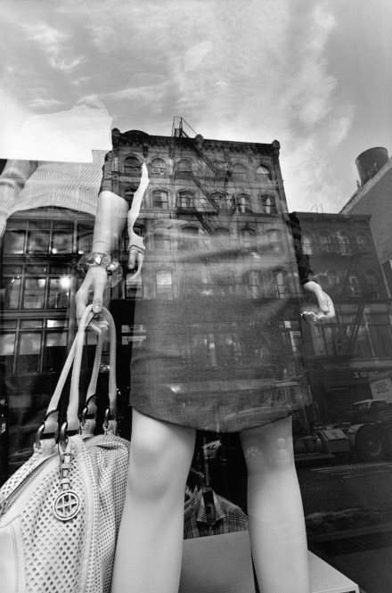 Black and white photograph of a mannequin in a storefront with the reflection of the buildings across the street in the glass window