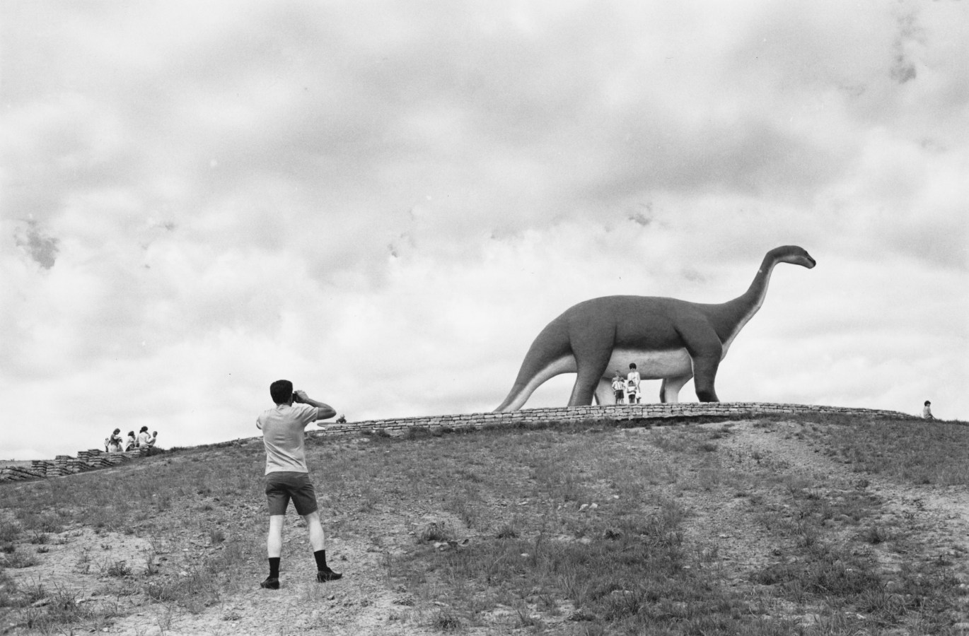 Black and white photograph of a person taking a family photograph in front of a life-size dinosaur statue