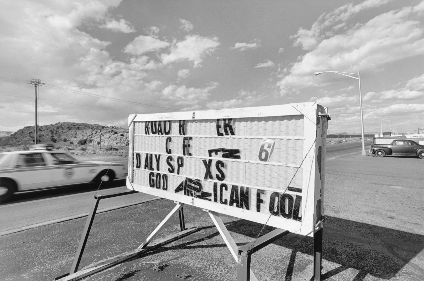 Black-and-white photograph of roadside sign missing letters with a police car speeding in the background and a cloudy sky