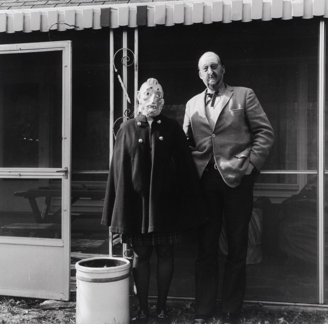 Black-and-white photograph of two people in rubber masks standing in front of a screened-in terrace