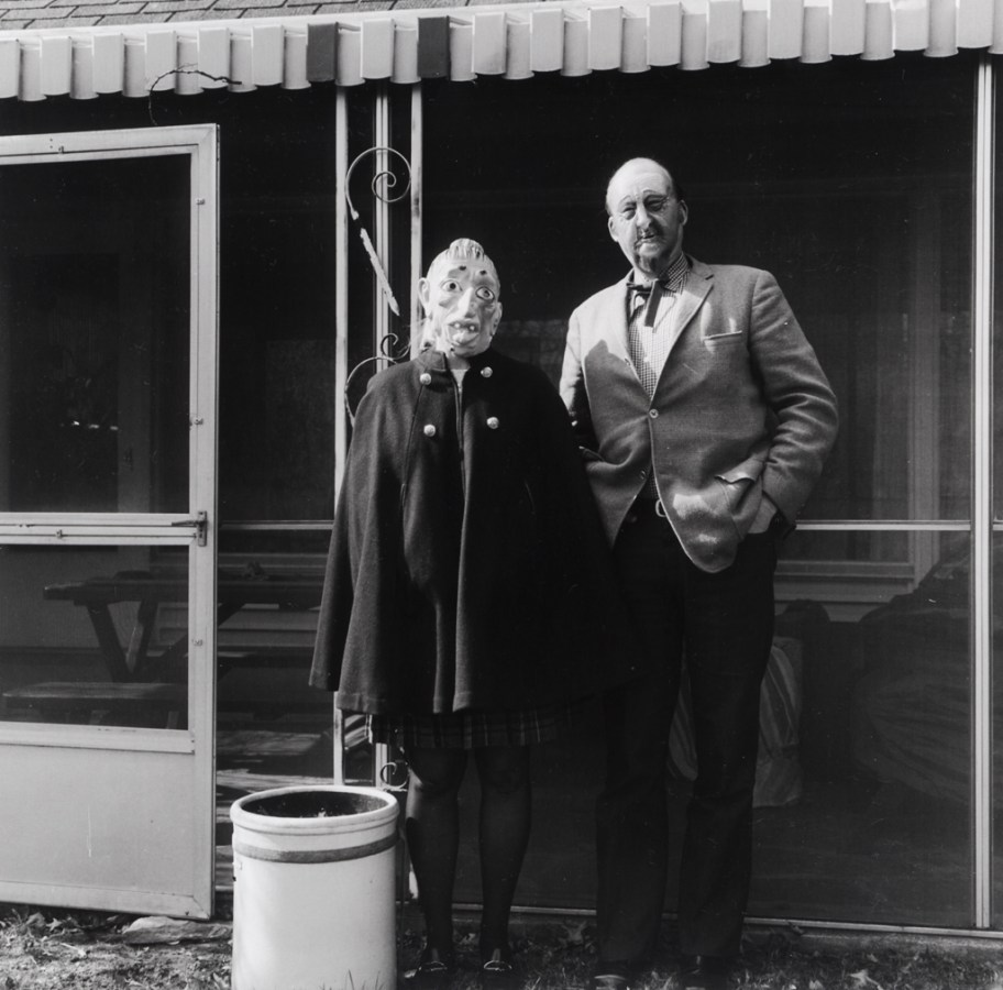 Black-and-white photograph of two people in rubber masks standing in front of a screened-in terrace