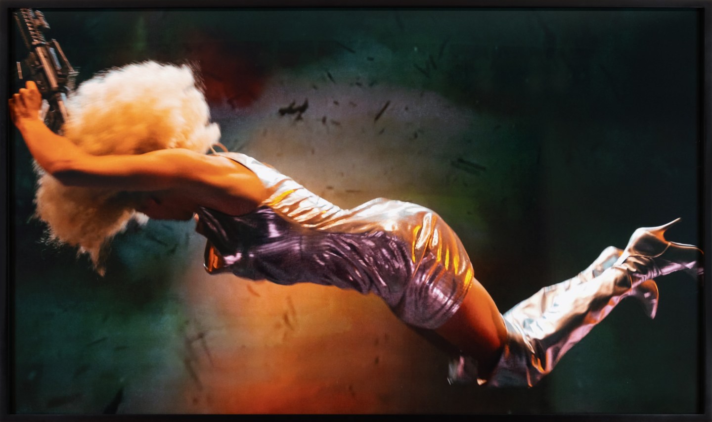 Color photograph depicting a woman holding a gun in midair with explosion behind framed in black frame