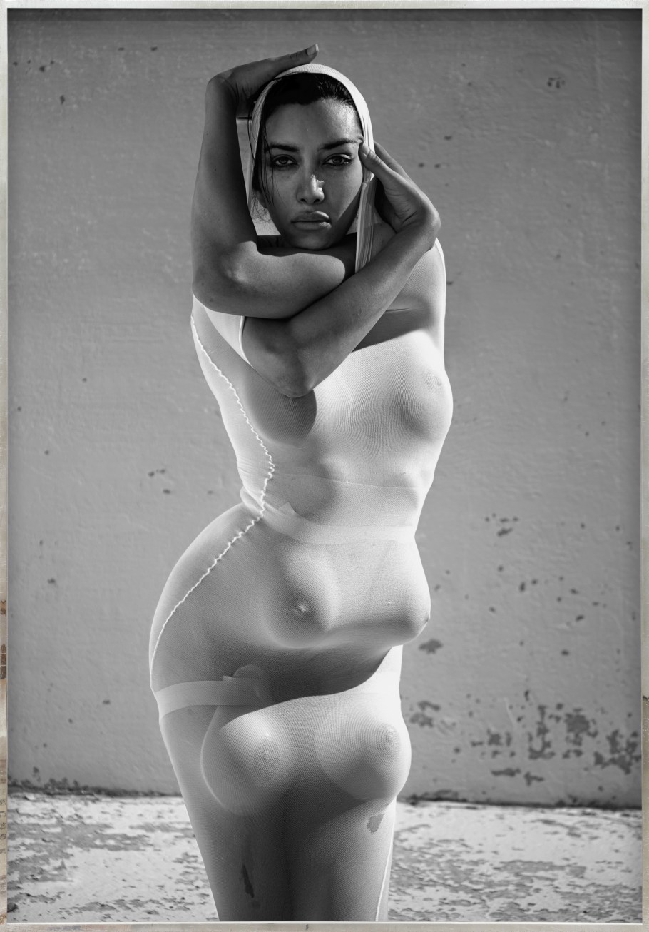 Color image of a black and white photograph of a figure clothed in white with multiple breasts and arms contorted around their head framed in distressed metal