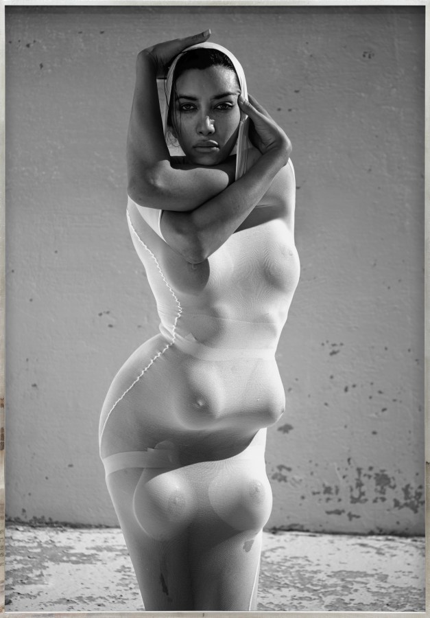 Color image of a black and white photograph of a figure clothed in white with multiple breasts and arms contorted around their head framed in distressed metal