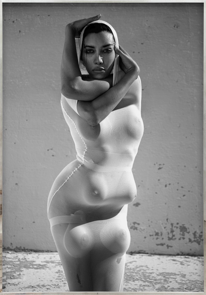 Color image of a black and white photograph of a figure clothed in white with multiple breasts and arms contorted around their head framed in distressed metal