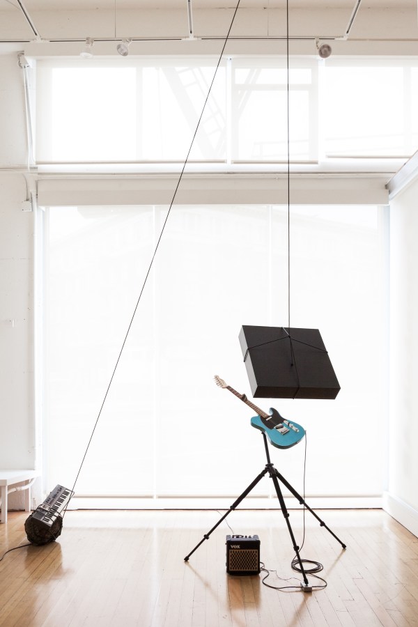 Color image of a mixed media sculpture consisting of an amp with a guitar and keyboard intwined with a cord attached to a suspended lightbox in a white walled gallery
