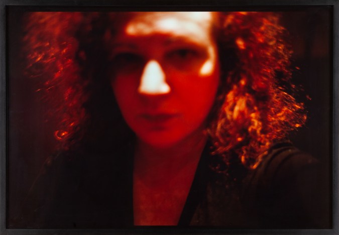 Color photograph of an out-of-focus woman's face framed by frizzy hair under orange light
